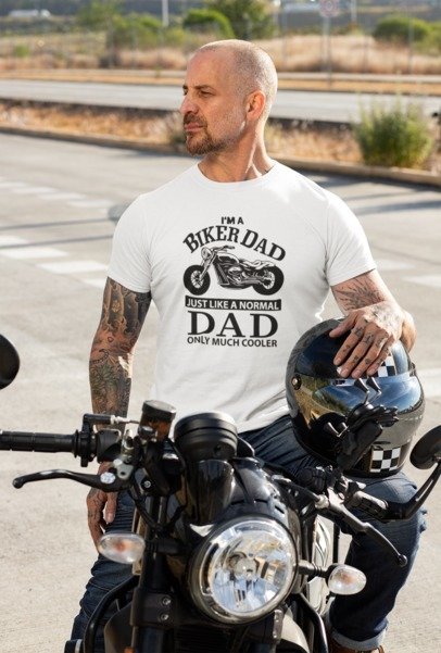 Man wearing a white I'm a Biker Dad graphic T-shirt beside a motorcycle, holding a helmet