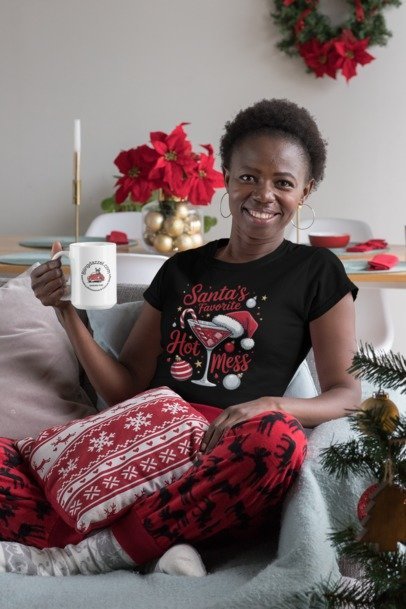 Model wearing a black Santa's Favorite Hot Mess graphic T-shirt and holding a mug beside Christmas decorations