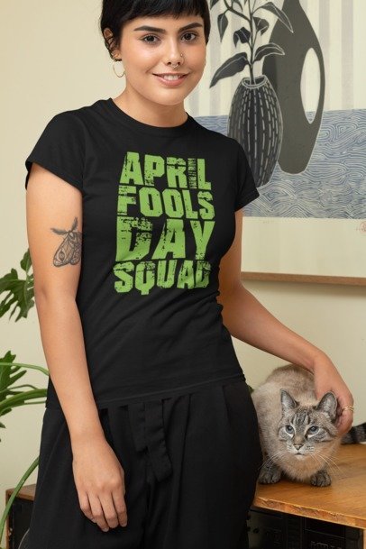 Model wearing a black April Fools Day Squad graphic T-shirt with green lettering beside a cat