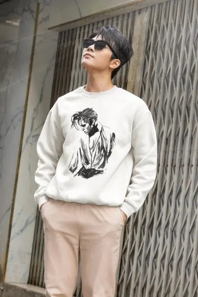 A man wearing a cream-colored crewneck sweatshirt with a black ink sketch of a person on the front, paired with light beige pants and black sunglasses, standing against a textured gray wall.