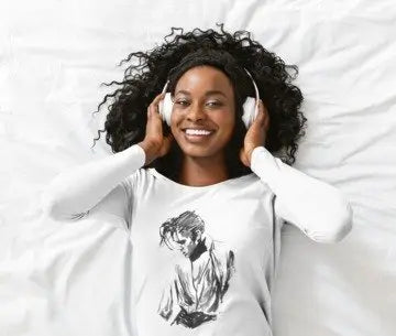 A smiling woman with curly black hair lies on white bedding, wearing white over-ear headphones and a white long-sleeve T-shirt with a black sketch-style graphic on the front.