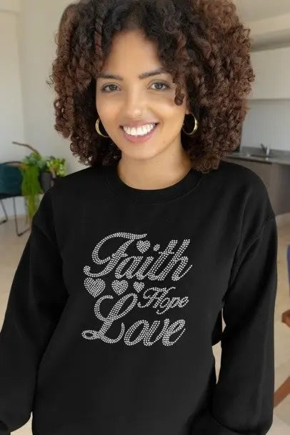 Person wearing a black sweatshirt with 'Faith Hope Love' text in rhinestones, indoors.