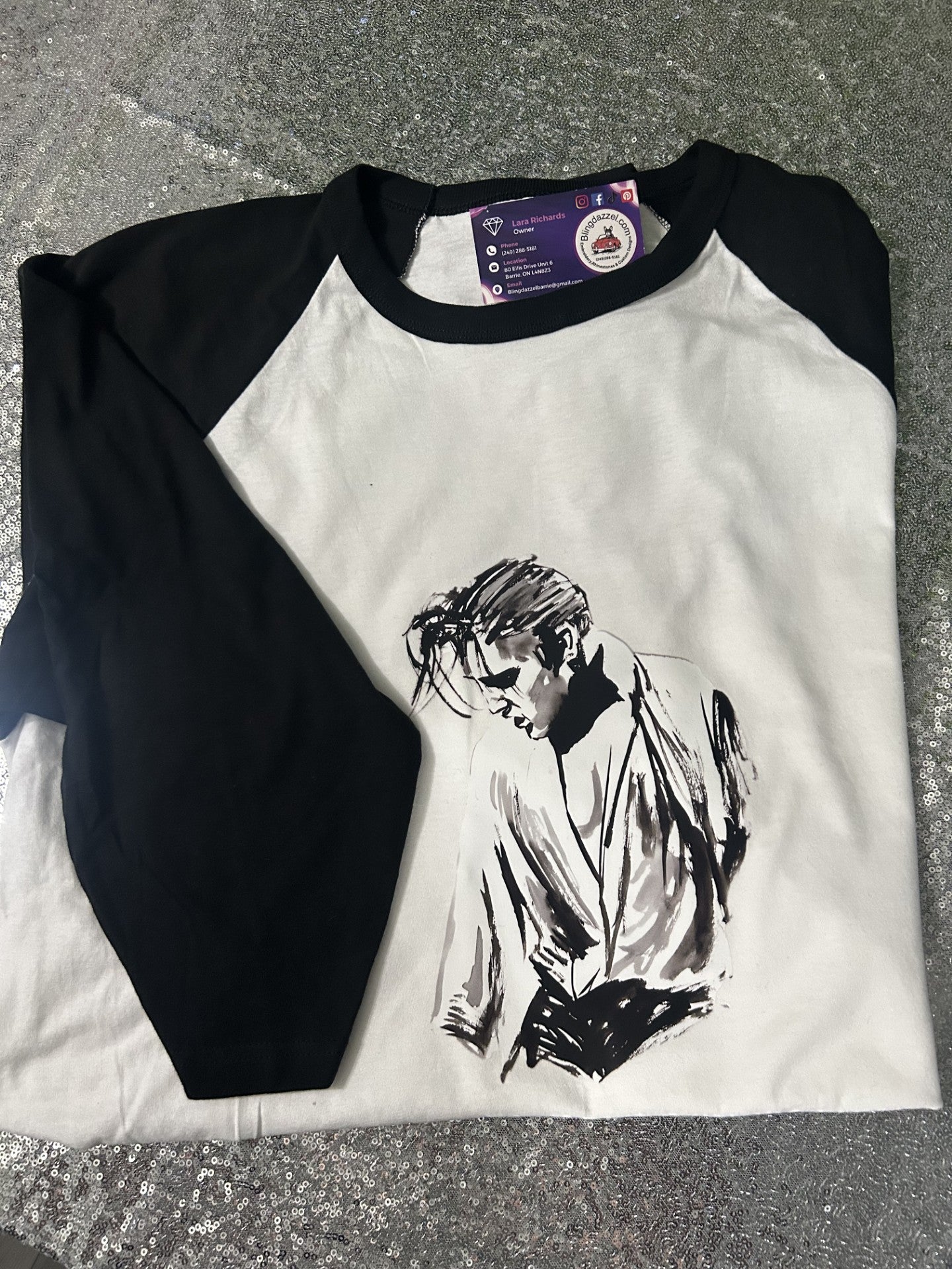 A black and white raglan baseball t-shirt with a black ink sketch of a person on the front, laid flat on a gray textured surface.