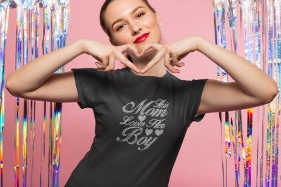 Person wearing a black t-shirt with 'This Mom Loves Her Boy' text, making heart shapes with hands against a colorful curtain background.